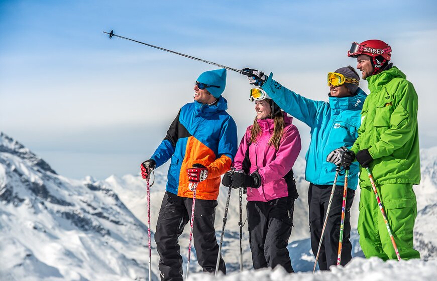 Three smiling skiers standing with Oxygene ski instructor who is pointing at something in the distance with his ski pole