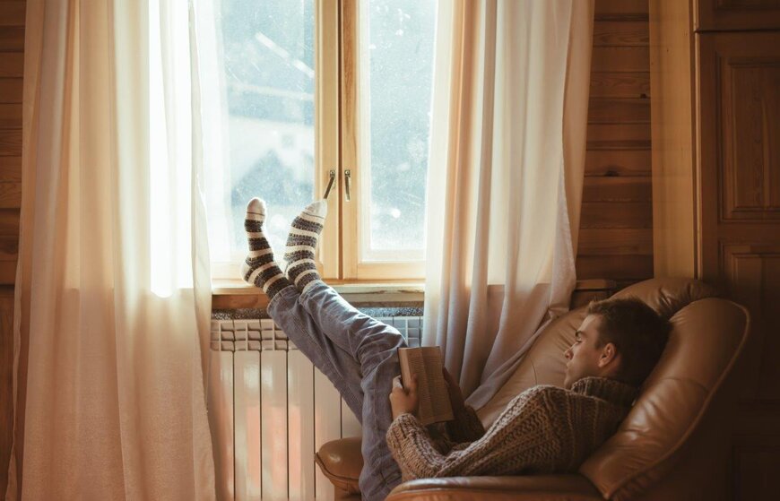 Young man reading book in cosy leather armchair by window with socked feet resting on window sill