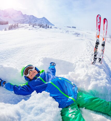 Smiling boy in ski kit doing snow angel in powder snow next to ski slope