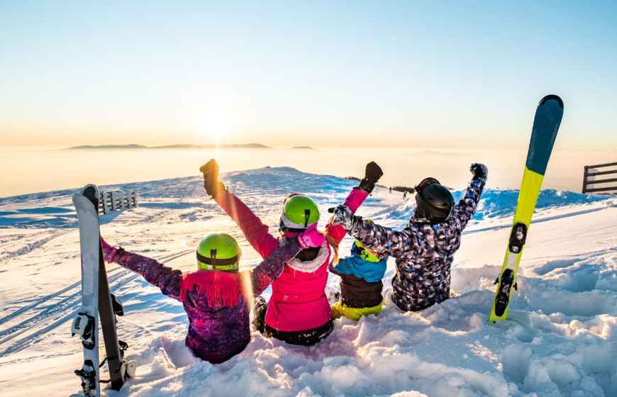 Family of four sitting in snow next to skis with hands in the air looking at the sun