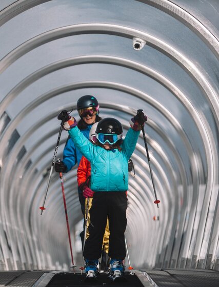 Child holding ski poles in the air whilst coming up covered magic carpet ski lift