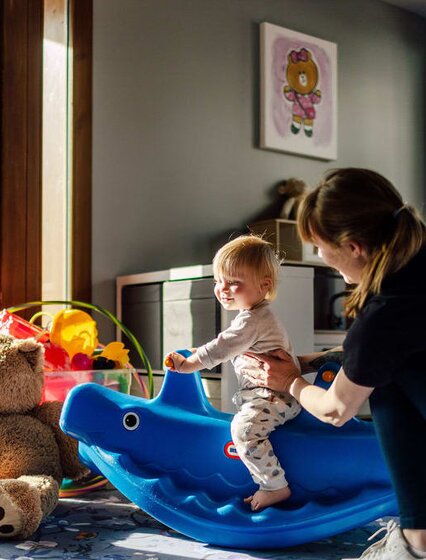 Blonde toddler playing on plastic ride on toy with ski nanny in Bear Cubs childcare