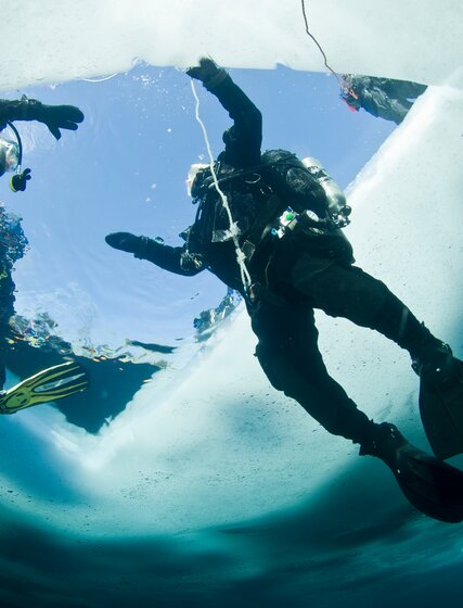 Two people in diving gear swimming below an ice hole