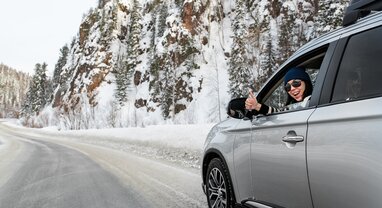 Smiling woman giving thumbs up from grey car on snowy mountain road
