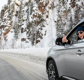 Smiling woman giving thumbs up from grey car on snowy mountain road