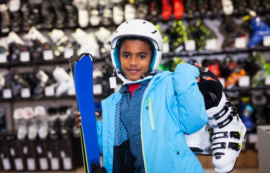 Young boy wearing ski jacket and helmet, holding skis and boots