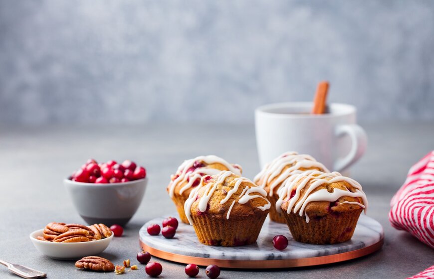 Four iced muffins, pecan nuts, cranberries and a hot chocolate in background