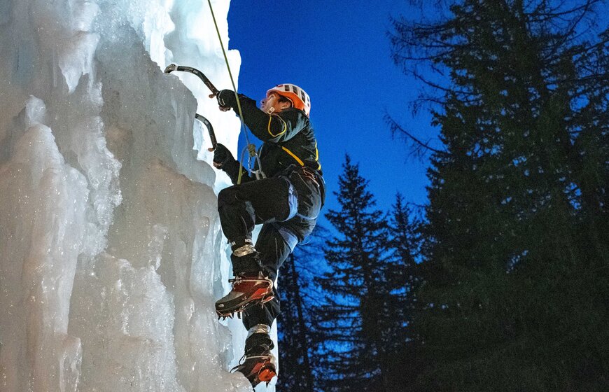 Man wearing helmet and crampons using two ice axes to climb floodlit ice tower in La Plagne 