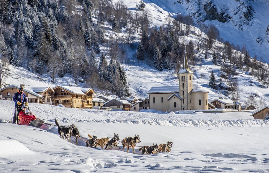 Mother and two children and musher being pulled by husky dogs through snow in La Plagne with church in background