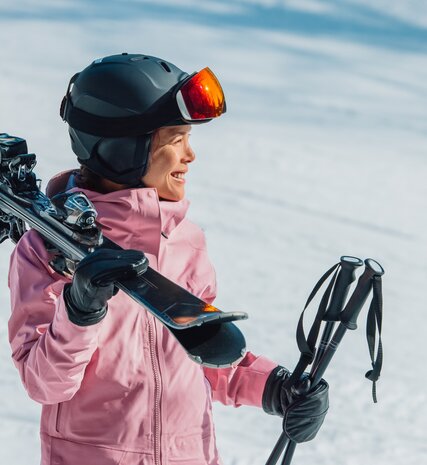 Smiling female skier in pink ski jacket and grey helmet carrying skis and poles