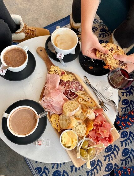 Charcuterie platter on small table wiht hot chocolates, glass of red wine and person holding cookie at Floe Bar in La Plagne